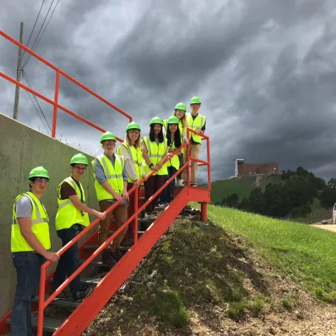Eight students stand on an outdoor stairway, with the Yates Headframe in the background