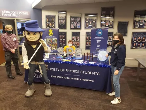 A professor, Mines Mascot, Sam Schleich and Serenity Engle gather around an SPS table