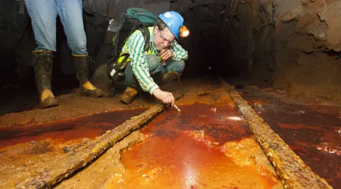 Dr. Dave Bergmann takes a sample from an underground area at SURF in between two train rails in rust colored fluid