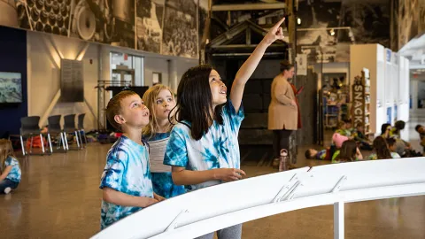 Students engage in STEM learning at the Sanford Lab Homestake Visitor Center in lead.  Three children are looking up at a model hanging from the celing. 
