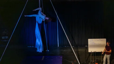Amber Sky Aerial performs "The Physics Behind Aerial Arts" which includes physicist Gavin Cox at a whiteboard and an aerial arts teacher, Kaitlyn Abrahamson on the silks, during the 2025 celebration of Neutrino Day in Lead, SD. 