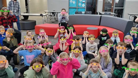 A group of students, wearing diffraction glasses, look on with expressions of amazement as light sources scatter into a kaliedoscope of rainbows.